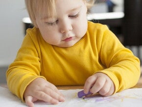 Bébé avec un pull jaune est assis à son bureau dans sa classe et est concentré à dessiner avec un crayola sur une feuille Bébé avec pull jaune assis à un bureau dessine
